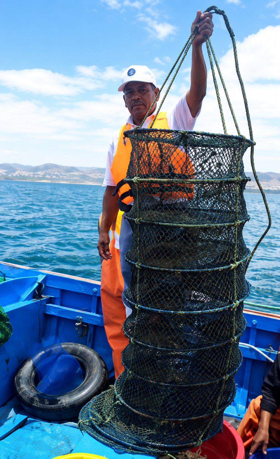 Pêcheur en gilet orange tenant une nasse sur un bateau, en mer
